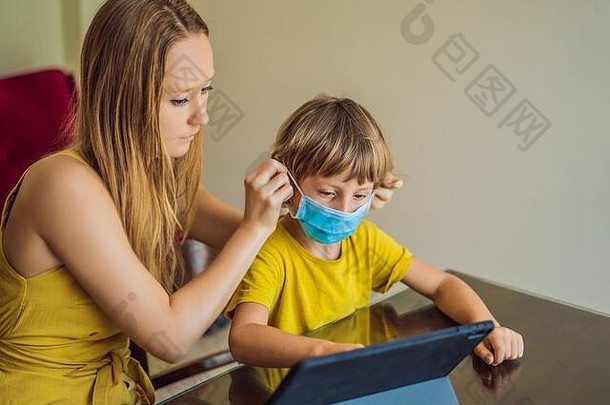 男孩在家里用平板电脑在线学习。妈妈帮助他学习。妈妈和儿子戴着医用口罩以防<strong>冠状病毒</strong>。学习期间