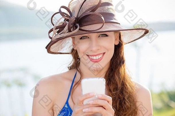 微笑的戴着帽子的美女用智能<strong>手机</strong>看着室外的摄像机，夏<strong>日</strong>公园的海湖背景，闲暇时间，正面的<strong>人类</strong>情感表情