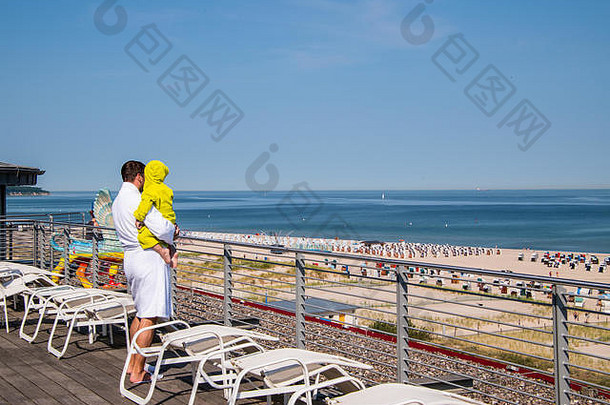 穿着白色长袍的男人和穿着绿色长袍的孩子在酒店露台上用<strong>多张</strong>躺椅眺望海滩和海洋