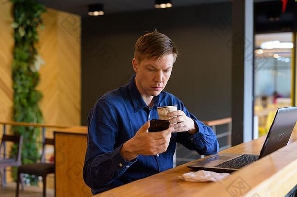 年轻英俊的商人，拿着咖啡，用手机，坐在咖啡馆里，远远的