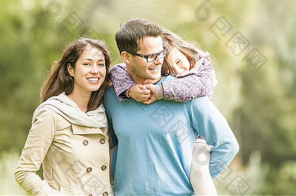 年轻快乐的三口之家一起在户外玩耍。漂亮的小女儿背着她父亲。父母和女孩看起来很高兴