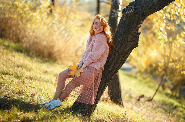 一个迷人的红发女孩的肖像，一张可爱的脸。穿着毛衣和珊瑚色裙子的女孩在秋天公园摆姿势。在一个女孩的手里