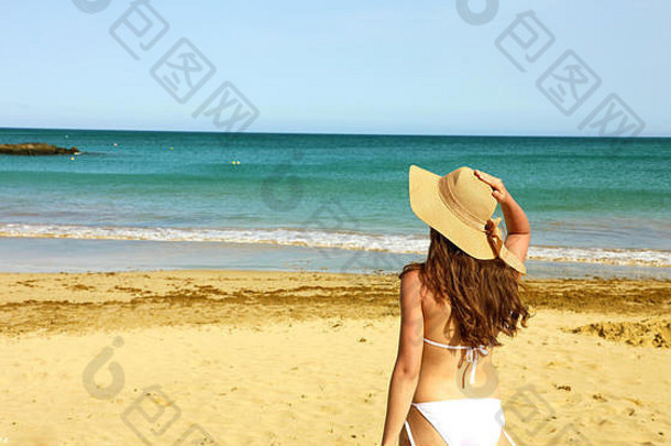 美丽的比基尼女士戴着草帽在兰扎罗特享受海风。在金丝雀般的夏日，年轻女子仰望大海的背影显得轻松<strong>自如</strong>