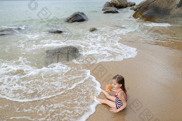 早上，小女孩和洋娃娃坐在海浪中的沙滩上。