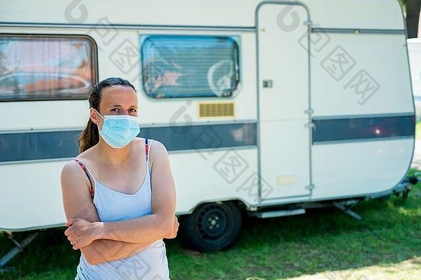 在一个度假胜地，一名戴着医用面罩的妇女站在露营拖车前。