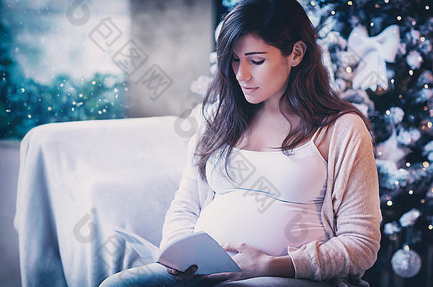 可爱温柔的孕妇坐在家里装饰精美的圣诞树旁看书