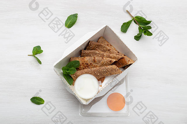 白色午餐盒里夹着酸奶油沙司的煎饼俯视图躺在绿色旁边的白色桌子上。饮食零食的概念。