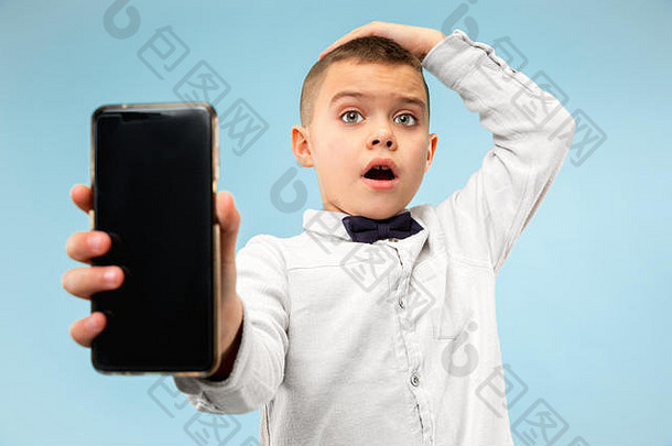 室内肖像有吸引力的年轻的男孩孤立的蓝色的背景持有空白智能手机微笑相机显示屏幕感觉快乐惊讶人类情绪面部表达式概念