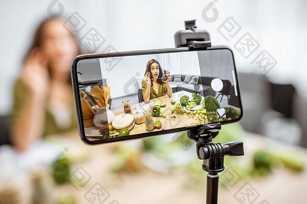 一名年轻女子在智能手机上录制关于健康饮食的视频日志。坐在家里有很多绿色素食食品配料的桌子旁，在手机屏幕上特写