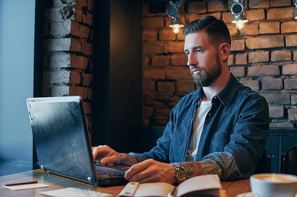 英俊的年轻人一边在咖啡馆喝咖啡一边用笔记本电脑工作