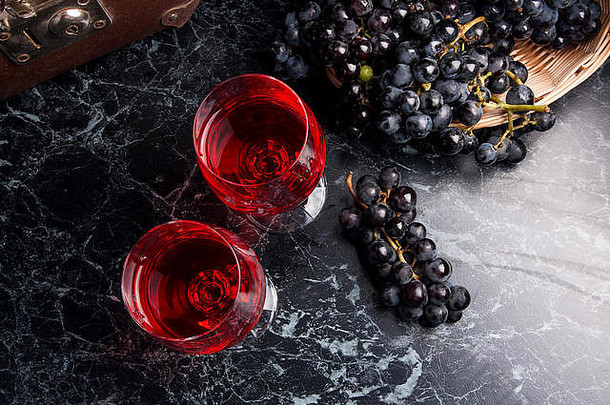 两杯红酒，深色大理石背景。深色大理石背景上的蓝色成熟葡萄。黄色篮子里的成熟葡萄和背面的旧箱子
