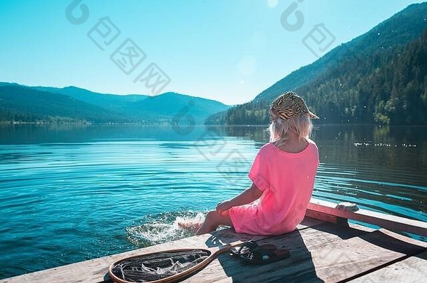 一个金发的年轻女孩坐在码头上，在阳光明媚的日子里欣赏湖景和周围的群山。
