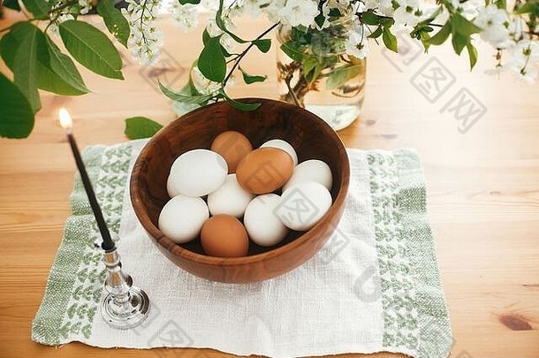 天然复活节彩蛋装在时尚的木制碗里，在樱桃花和绿叶的背景下，用亚麻布制成的复古蜡烛装饰。复活节快乐概念。