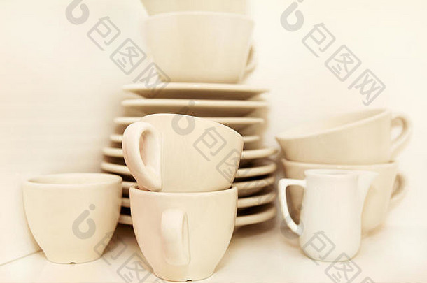 小壶和白色咖啡杯，白色背景的架子上有服务盘。咖啡吧里有带托盘的咖啡杯。白色陶器套装。