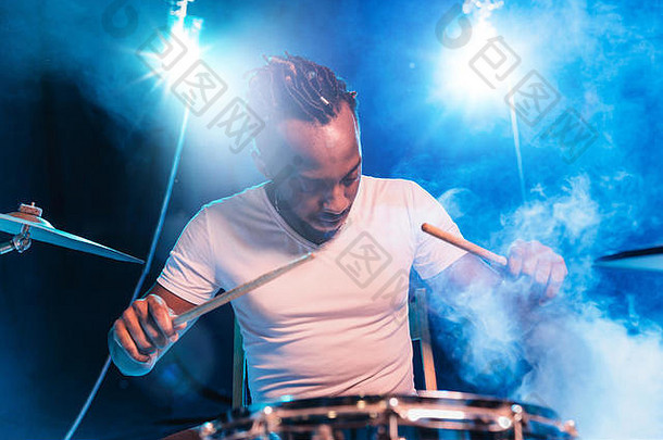 年轻的非裔美国爵士乐音乐家或鼓手，在蓝色工作室背景下，在他周围的炽热烟雾中打鼓。音乐的概念，爱好，灵感。快乐迷人的艺术家的彩色肖像。