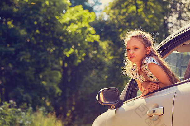 在夏日明亮的绿色自然背景下，开着嘴从车窗向外张望，开心快乐地享受旅行的小女孩。特写夏日肖像。L