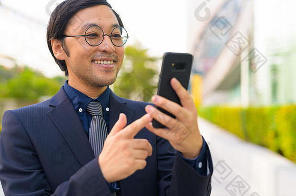 快乐的亚洲商人在城市户外使用手机的脸