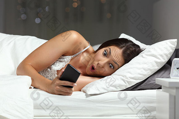 惊讶的女孩在家里晚上躺在床上的智能手机里发现了惊人的在线内容
