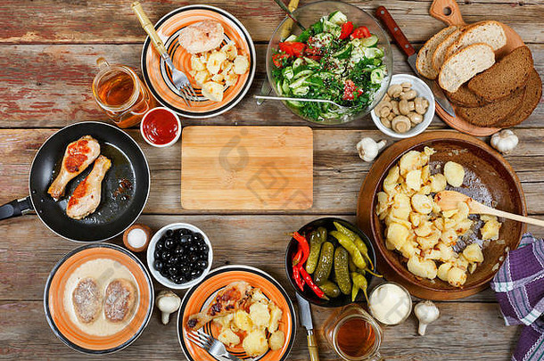 餐桌上有各种各样的菜肴，烤鸡腿，煎锅里有蔬菜的炸土豆，木桌上的沙拉和零食，俯视图