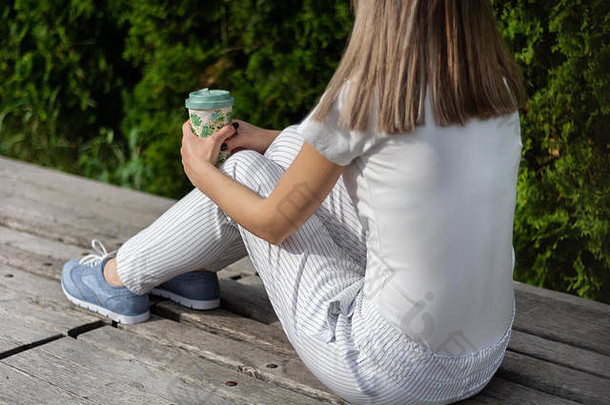 穿着条纹裤坐在长凳上，手里拿着一杯咖啡的时髦女孩。天气好时在公园休息的女人。春夏服装流行