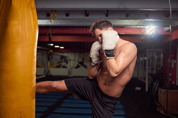 一个年轻的赤膊男子，拳击手打拳击袋，在室内健身房练习，戴拳击手套，上身射击。