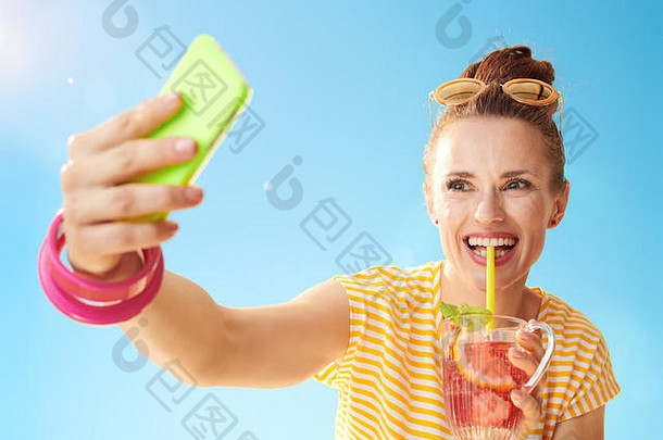 微笑健康的女人黄色的衬衫蓝色的天空让人耳目一新鸡尾酒采取自拍智能手机