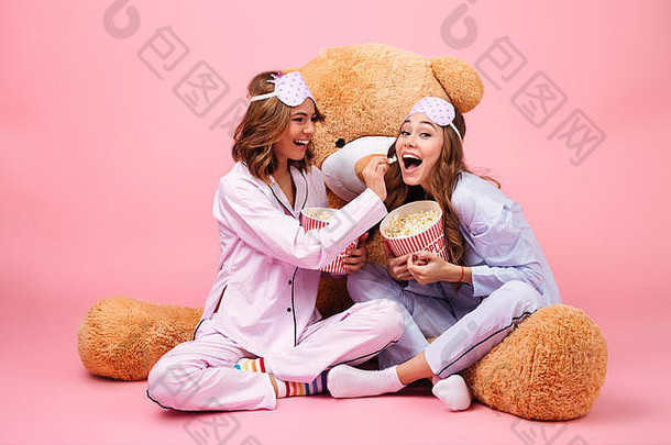 两个穿着睡衣、兴高采烈的漂亮女孩和一只大泰迪熊坐在一起，吃着粉色背景下的爆米花