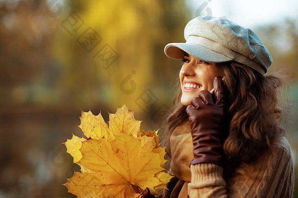 你好，秋天。穿着毛衣、帽子、手套和黄叶围巾的快乐优雅的中年妇女坐在外面的长椅上，用智能手机