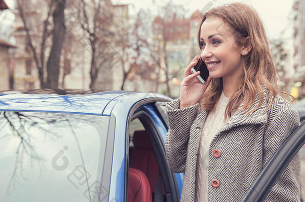 穿着外套的漂亮女人站在汽车旁的街道上微笑着打电话。