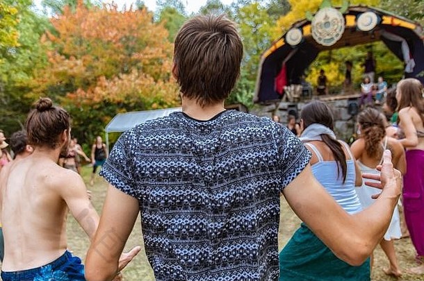 从后面可以看到一个20多岁的波西米亚男孩，蓬乱的棕色头发和蓝色印花t<strong>恤</strong>，在<strong>节</strong>日期间与模糊的人在背景中跳舞