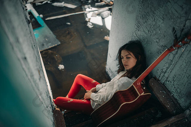 一位年轻女子穿着白色衬衫和红色紧身衣，手拿吉他坐在废弃的旧船里。