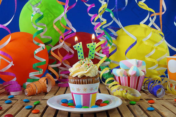11生日纸杯蛋糕，蜡烛在乡村木桌上燃烧，背景是五颜六色的气球，背景是蓝色墙上的塑料杯糖果