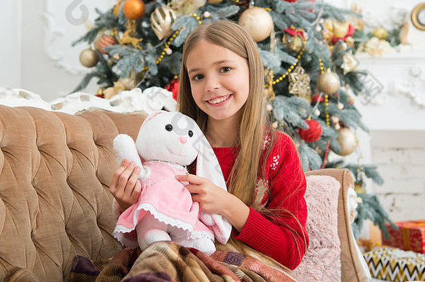 它融化了我的心。小孩玩软玩具。小女孩和可爱的兔子在圣诞树旁。小女孩抱着兔子玩具。小孩带着礼物开心地笑着。最好的圣诞玩具。