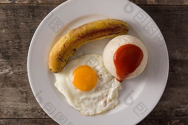 Arroz是一种典型的古巴米饭，在木桌上的盘子里放着炸香蕉和煎蛋。顶视图。