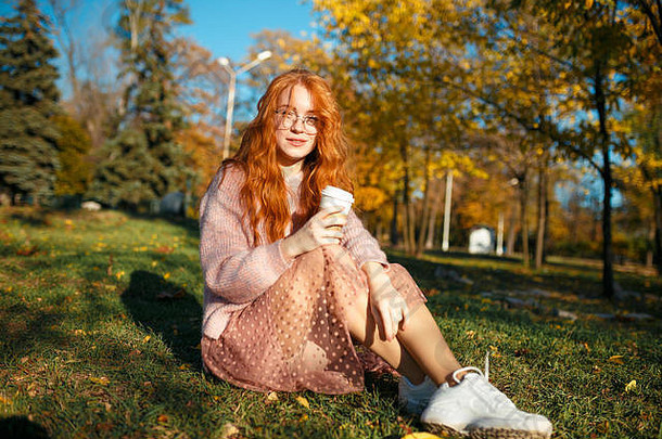 一个迷人的红发女孩的肖像，戴着眼镜，一张漂亮的脸。一个穿着毛衣和珊瑚色裙子的女孩在秋天公园摆姿势。在少女时代