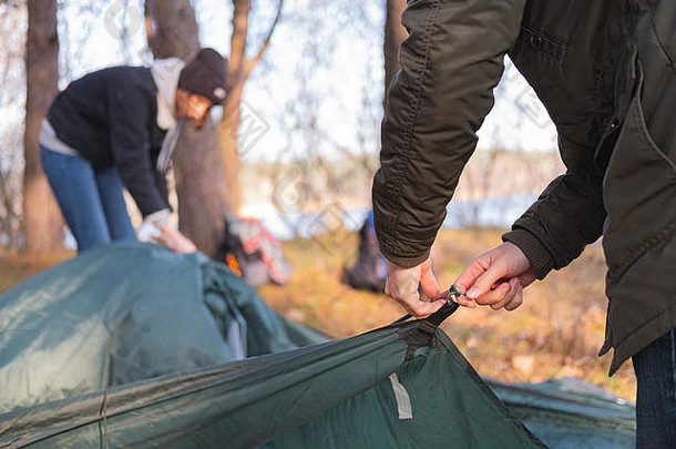 两个穿着暖和衣服的人搭起帐篷。寒冷季节徒步旅行：男人和女人在河边搭起帐篷