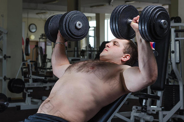 在健身房里，一个<strong>肌肉</strong>发达的年轻人在倾斜的长凳上做哑铃式胸部按压