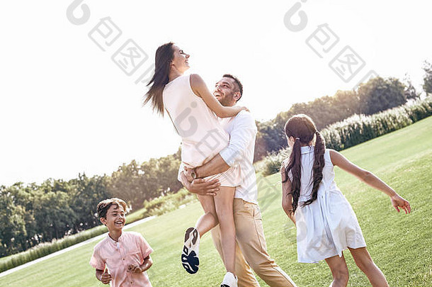 结合。一家四口走在草地上，丈夫抱着妻子，开心地拥抱着，孩子们在奔跑