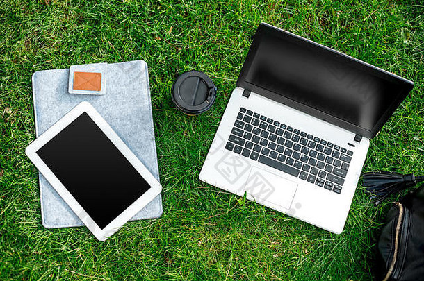户外公园绿草上的笔记本电脑，配有咖啡杯、咖啡袋和平板电脑