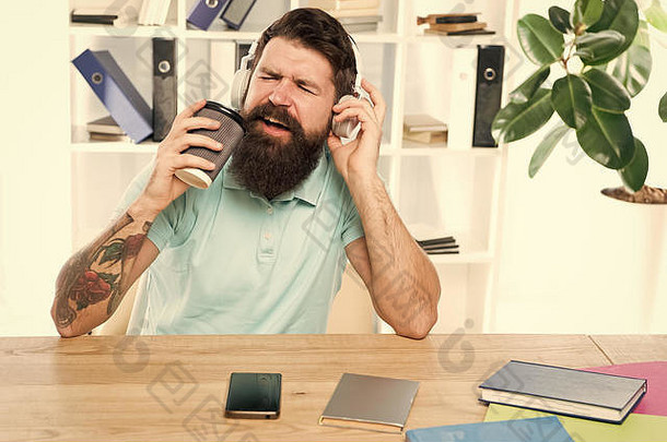 正常工作日。留胡子<strong>的</strong>男人戴着耳机坐在办公室听音乐唱歌。拿着咖啡杯<strong>的</strong>工人用最喜欢<strong>的歌曲</strong>开始工作日。充满灵感和活力。办公室生活<strong>节奏</strong>。