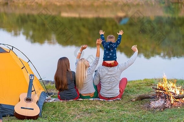 从后面。一群快乐的朋友和孩子肩并肩在河边露营，手挽着手跳舞，欣赏风景。家庭假期很有趣。