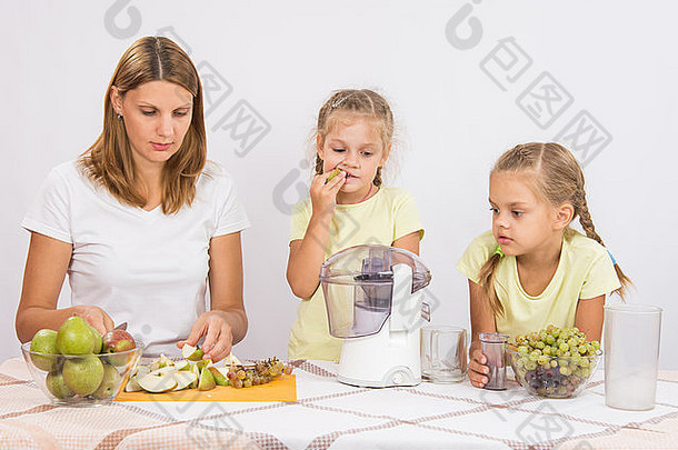 年轻漂亮的母亲和两个女儿坐在桌旁，用榨汁机从梨和葡萄中榨汁