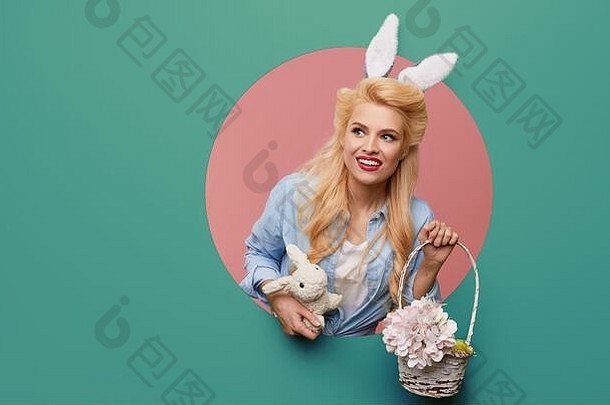 一个戴着复活节兔子耳朵、拿着一篮彩蛋的年轻女人从墙上的一个圆孔爬了出来。薄荷醇或绿松石色背景