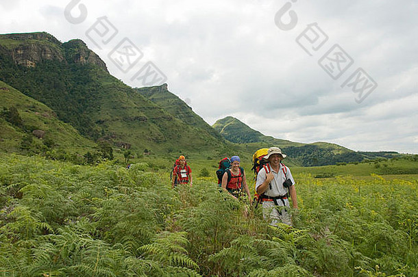 三名徒步旅行者背着背包沿着一条小径行走，小径两旁是蕨类植物、悬崖和山丘，后面是阴云密布的天空。