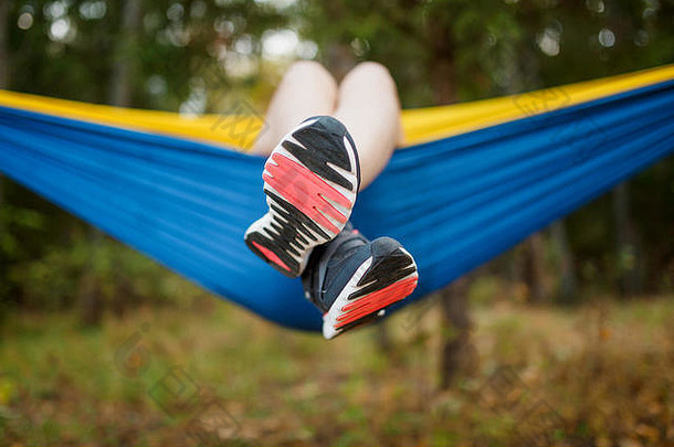 照片中的女孩穿着运动鞋，双腿悬垂，躺在树林中的吊床上