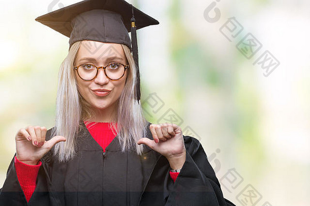 年轻的金发女郎穿着毕业制服，在孤立的背景下，脸上带着自信的微笑，用手指指着自己，骄傲而快乐