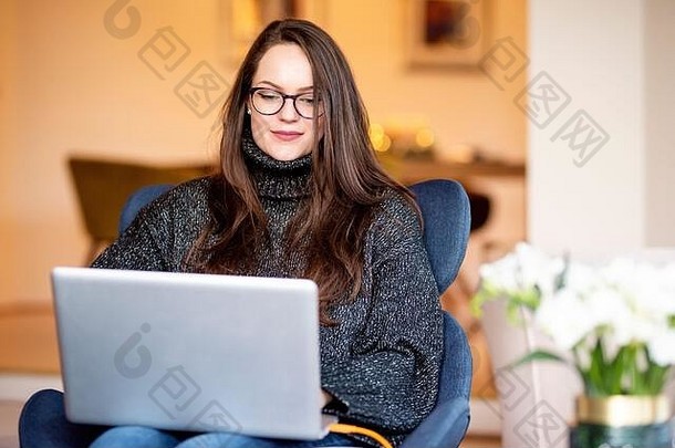 这张照片拍摄的是一位微笑的年轻女子坐在椅子上，在家里工作时使用笔记本电脑。