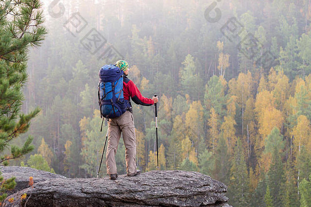 黎明时分，在班达纳，背着背包和登山竿的男子站在秋林背景的岩石上。