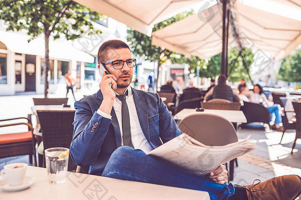 戴眼镜的年轻商人坐在咖啡馆里看报纸，打<strong>手机</strong>。