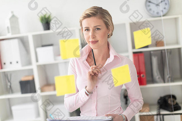 一个年轻的金发女孩站在办公室里，旁边是一块透明的贴纸板，手里拿着文件和铅笔。